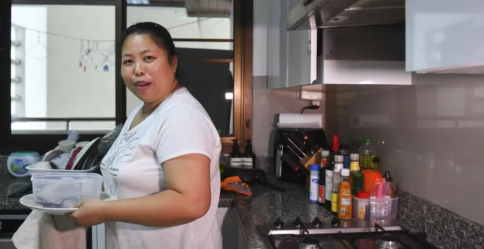 The image shows a woman standing in a kitchen holding a plastic container and a plate, surrounded by various kitchen items and condiments on the countertop.