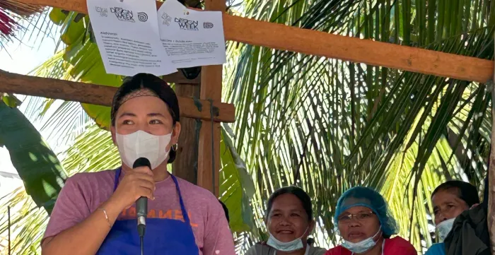 Jonalyn is wearing a blue apron and speaking over a microphone.