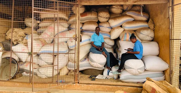 Kalian Sampoa Gumah conducts a survey with a male farmer. They are seated on large white bags of grain in a grain storage shed. (Ghana, 2025)