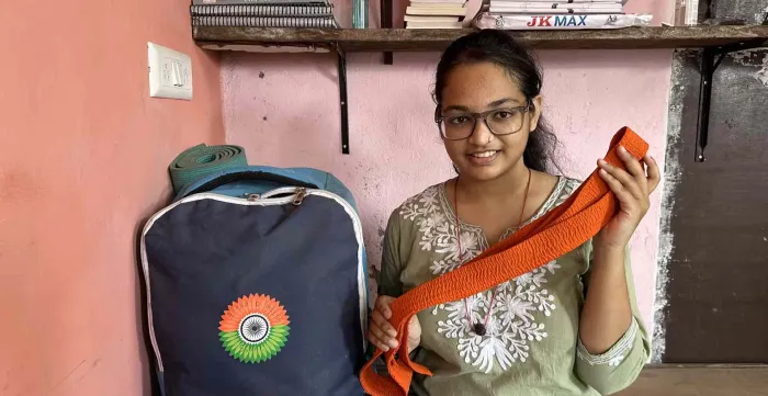 Reena est assise sur son lit et regarde la caméra. Derrière elle se trouve une étagère avec des livres. Dans ses mains, elle tient sa ceinture de karaté orange. Surat, Inde. (2024)