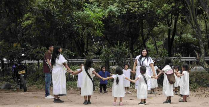 Yoraima Cristina Navarro se trouve au sein d’un cercle composé d’enfants et d’adultes se tenant la main. Ils sont à l’extérieur. On aperçoit des arbres au feuillage verdoyant. (2025)