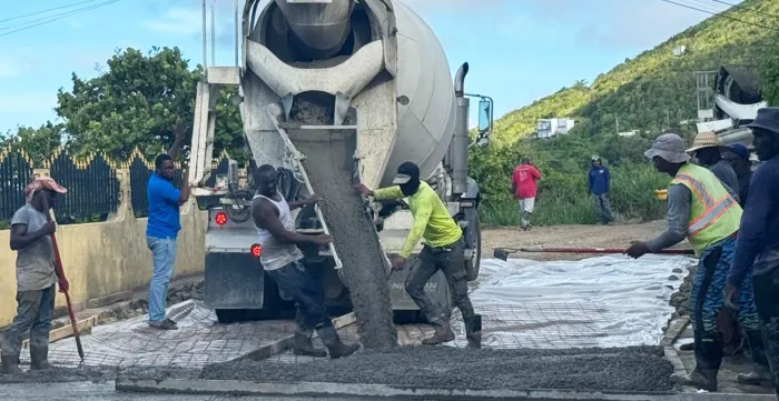 A group of construction workers is actively pouring and spreading concrete from a cement mixer truck on a road section. 