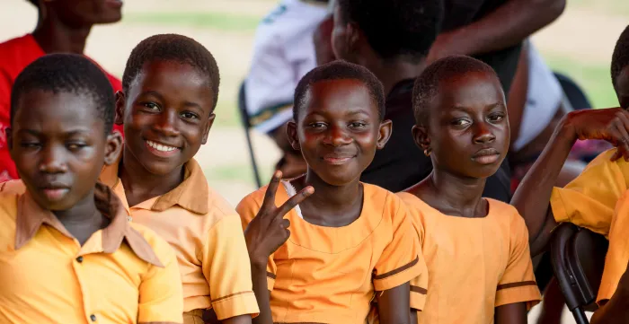 A group of children in yellow school uniforms sit and smile at the camera during a visit by the Japanese Ambassador to their school in Beposo, Ghana. One child flashes a peace sign, while others look on. The visit was part of the Climate Cocoa Project's support to child labour-free zones.