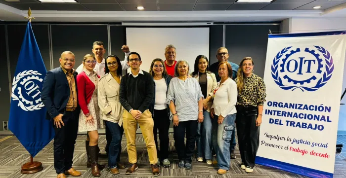 Grupo de doce personas posando en una sala de conferencias junto a una bandera y un cartel de la Organización Internacional del Trabajo (OIT). El cartel muestra el logotipo de la OIT y el lema ‘Impulsar la justicia social. Promover el trabajo decente’. Las personas están sonrientes