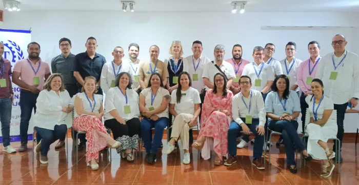 Aproximadamente 25 personas posa para una fotografía grupal en el interior de una sala iluminada con luz blanca. La mayoría lleva colgadas credenciales azules y verdes, lo que sugiere que asisten a un evento o taller. En la primera fila, algunas personas están sentadas y sonríen hacia la cámara; detrás, el resto del grupo está de pie. Al fondo se distingue una pared blanca y un cartel parcialmente visible con un logotipo de la OIT