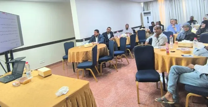 Business representatives at workshop, resource person speaks to them, with his laptop open and a big scream in the background 