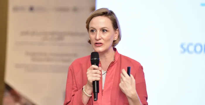A woman in a pink jacket speaks into a microphone while holding a pen, standing in front of a blurred presentation screen and banner.