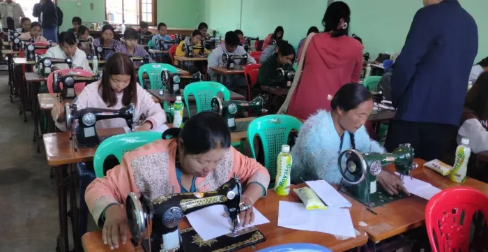 Women are practising with sewing machines in the training and a woman & a man are checking them.