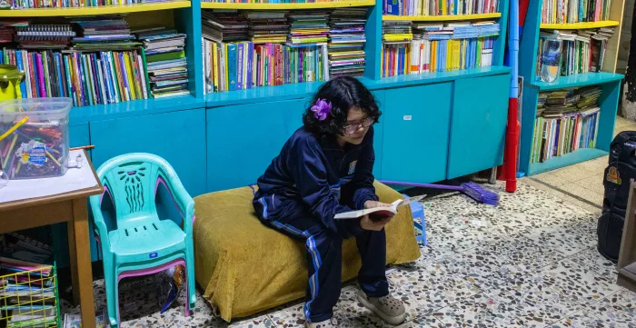 A teenage girl sits on a small bench inside a neighbourhood library, reading a book. Shelves filled with books line the wall behind her.