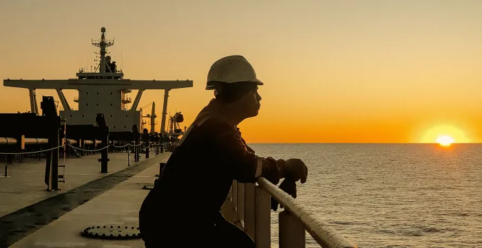 Seafarer standing at the railing of a cargo vessel, gazing at the horizon as the sun sets over the ocean