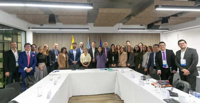 Grupo de alrededor de veinte personas en una sala de reuniones moderna, posando para una foto alrededor de una mesa en forma de U cubierta con manteles blancos. La mayoría viste ropa formal o de negocios. Al fondo se observan banderas institucionales, incluyendo la de Colombia y la de Naciones Unidas.