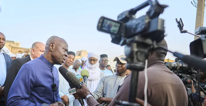 Le Directeur général de l’OIT, Gilbert F. Houngbo, s’exprime devant des journalistes lors d’une interview, avec des microphones et des caméras visibles, tandis que des responsables et des personnes présentes se tiennent derrière lui.