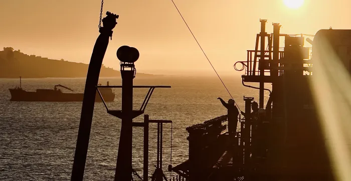 A lone seafarer stands on the deck of a ship in the sunset