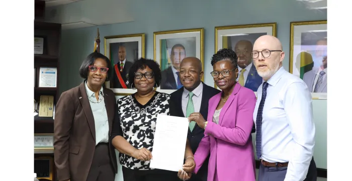 Alt Text: Signing of the MOU between ILO and JHWU  A professional group portrait features five individuals standing together in an office setting, holding a signed document. The group consists of three women and two men, all dressed in formal business attire.