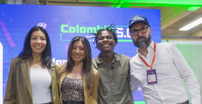 Una fotografía de cuatro personas sonriendo en un evento tecnológico. Están de pie una al lado de la otra frente a una gran pantalla azul que dice "Colombia 5.0" en verde y blanco.