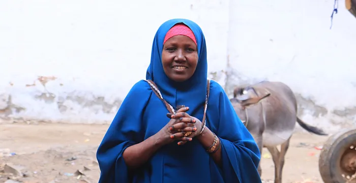 Portrait de Hawa Ali Mohamed. Elle se tient debout, les mains jointes, et porte un hijab et une abaya bleus. Une vache se tient derrière elle à l’arrière-plan.