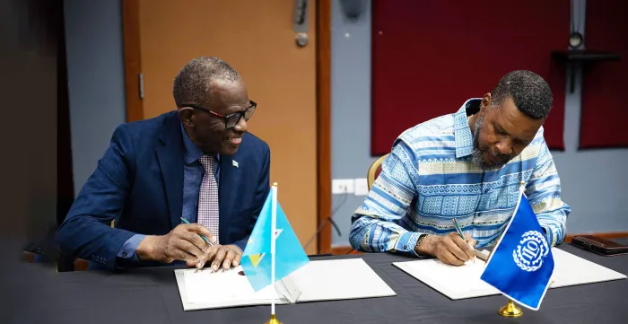 Two men seated at a table sign official documents, with the flags of Saint Lucia and the International Labour Organization (ILO) displayed in the foreground.