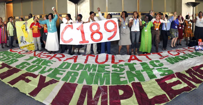 Group of domestic workers, trade union representatives and supporters standing together at the International Labour Conference, holding a large "C189" sign and a banner calling for ratification and implementation of the Domestic Workers Convention. Participants raise joined hands in celebration following the adoption of Convention No. 189 on decent work for domestic workers.