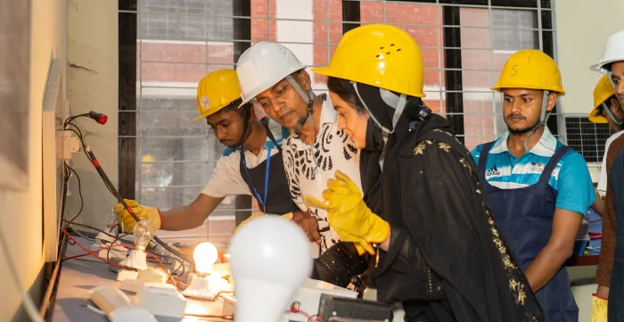Le directeur du centre de formation technique et professionnelle, Md. Ziaur Rahman, et quatre stagiaires, dont une jeune femme au premier plan, portent des casques et des gants de sécurité et observent une installation électrique équipée de deux ampoules allumées.
