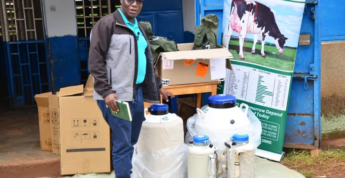 An artificial insemination specialist stands outside a cooperative dairy facility in Kayunga, Uganda, demonstrating livestock breeding equipment to farmers. Several insulated storage containers and transport canisters used for artificial insemination are displayed alongside packaging boxes, while an educational banner showing dairy cattle genetics and trait information stands in the background.