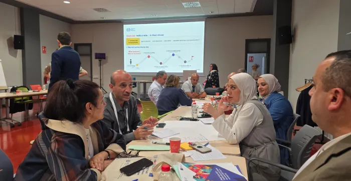 Participants seated around tables engage in small-group discussions during an interactive workshop session. Training materials, notebooks and documents are spread across the tables, while a presentation on youth pathways and service delivery is displayed on a screen in the background.