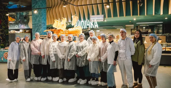A group of students wearing protective gowns, hairnets and shoe covers stand inside a bakery production facility alongside bakery staff.