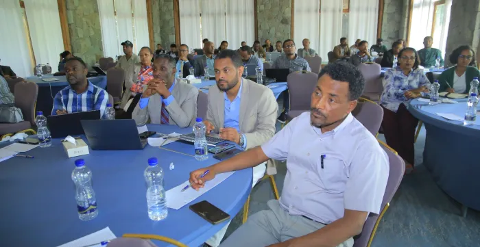 Participants seated around tables during a workshop session, listening and taking notes.