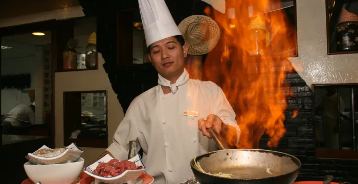 A chef wearing a white uniform and tall chef's hat cooks over a flaming wok in a restaurant kitchen.