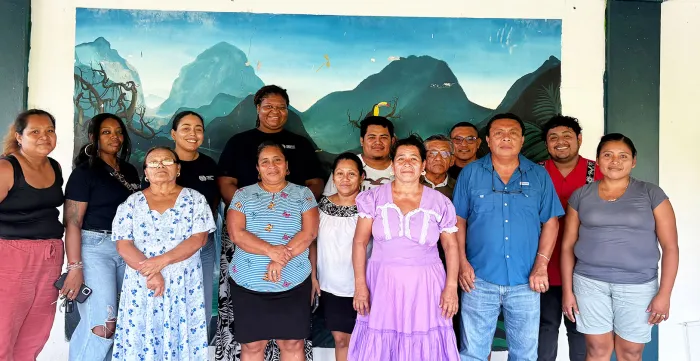 A group photo of thirteen individuals standing side-by-side outdoors against a painted mural backdrop depicting lush green mountains and a blue sky.