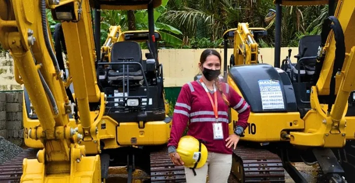 A woman wearing a high-visibility work shirt, face mask, and identification badge stands between two mini excavators while holding a yellow safety helmet.