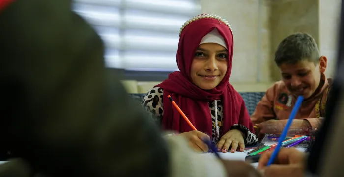 Yara wears a burgundy headscarf smiles while drawing with colored pencils during a classroom activity.