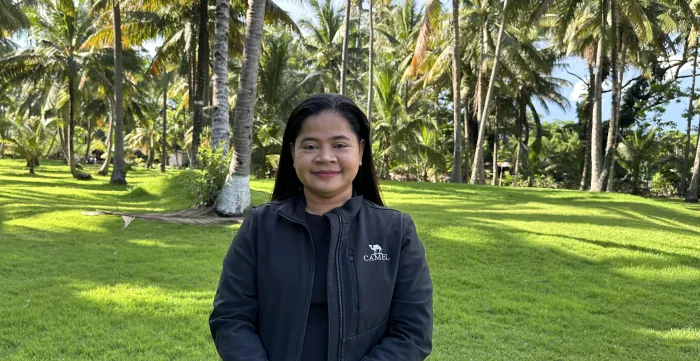 Melba wearing a black jacket and standing behind a green field with coconut trees