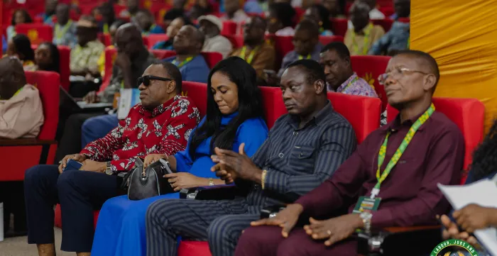 Several conference participants sit in a conference hall, listening to presentations. The audience includes men and women wearing accreditation lanyards during a labour conference in Ghana
