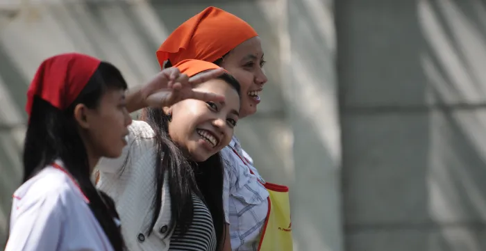 Three young women wearing orange headscarves standing together outdoors, smiling. One makes a peace sign while the others look ahead.
