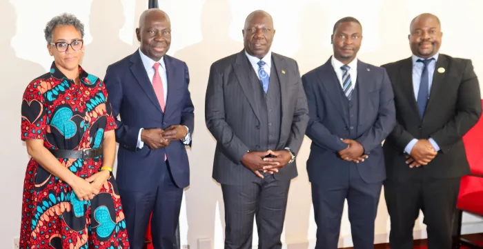 Five officials stand together for a group photo in an office. Four men wear dark business suits and one woman wears a patterned red, black and turquoise dress. They face the camera in front of a national flag and a framed portrait.