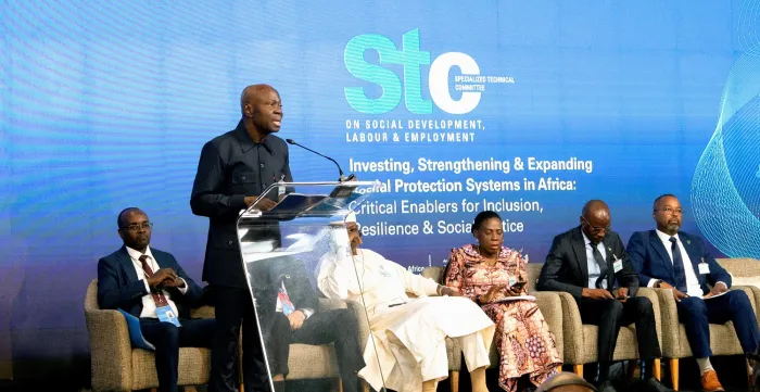 Smartly dressed black middle aged man speaking at a podium with other 6 black men and women in local dress and suits, sat behind him.
