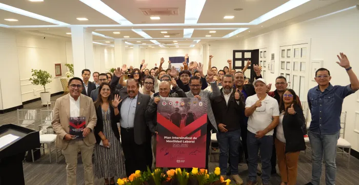 Grupo de personas posan en un salón de eventos y saludan con una mano levantado; mientras las personas en el centro del grupo sostienen un cartel sobre el Plan Intersindical de Movilidad Laboral 2025-2030