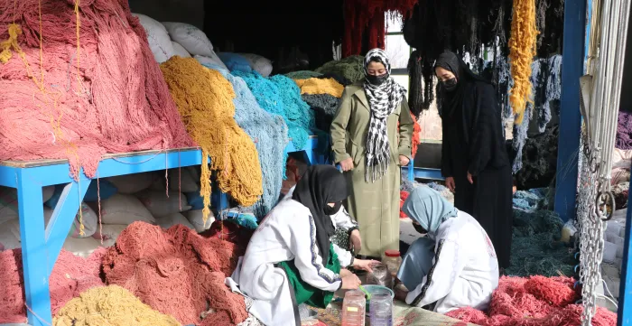 Women work among large bundles of colourful yarn while two other women observe their work.