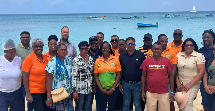 A group of fisheries knowledge-exchange participants standing together on a sandy beach, with bright blue ocean waters and small fishing boats behind them.