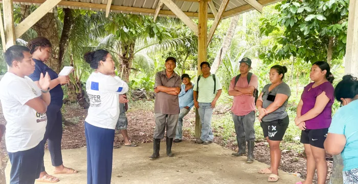A group of community members gathers under an open-air wooden shelter for a discussion led by a facilitator wearing an EU-ILO PROSPER long-sleeve shirt.