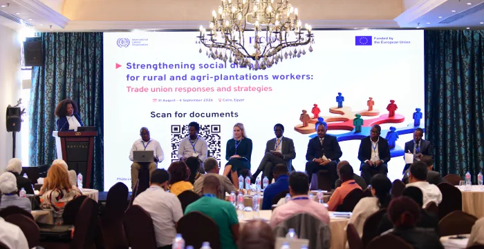 A speaker addresses participants seated in a conference room, with six panellists seated on stage in front of the event screen.