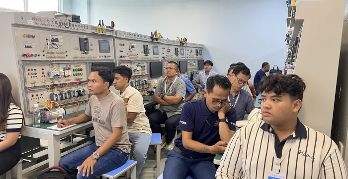 Several people sit at workbenches in a technical classroom, with shelves of electronic equipment behind them.