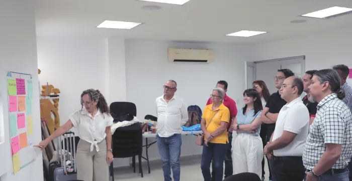 Grupo de personas compuesto por 4 mujeres y 7 hombres observan la cartelera realizada en una pared, que está siendo explicada por una mujer