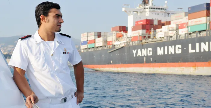 A coastguard officer in a white uniform stands aboard a vessel, with a large container ship passing behind him in the Port of Genoa, Italy.