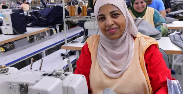 A garment worker sits at a sewing machine on the production floor of Eroğlu Manufacturing Studio in Ismailia, Egypt, with other workers at sewing stations in the background.