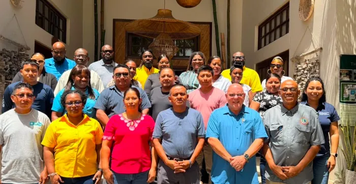 A large group photograph of approximately 25 to 30 people posing outside the entrance of a building. The diverse group of men and women are dressed in casual and business-casual attire, including brightly coloured t-shirts, polo shirts, and button-downs, and are organized in multiple rows facing the camera.