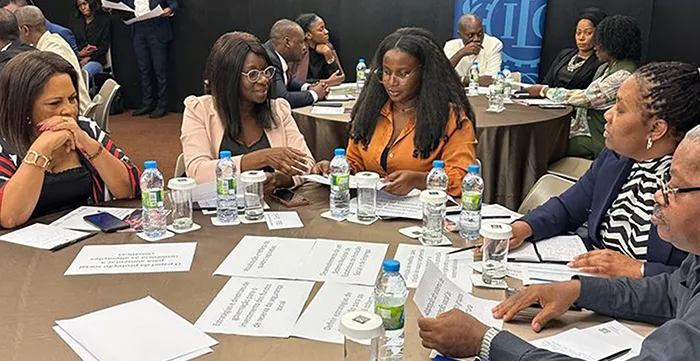 Grupo de pessoas conversam sentados a uma mesa. Na mesa estão papéis, garrafas de água e copos.  