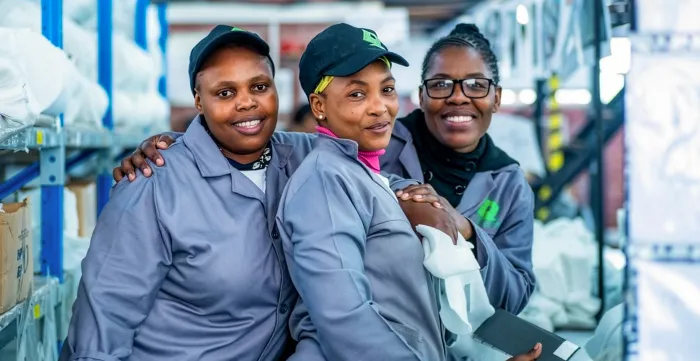 Three women factory workers in grey uniforms smile together during an ILO project field visit in South Africa in 2023.