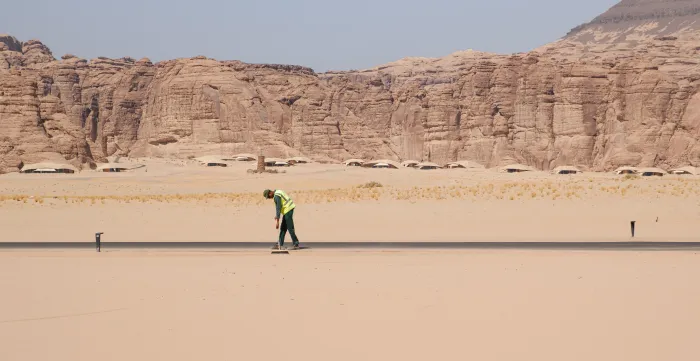 A worker wearing a high-visibility vest sweeps a paved road in a vast desert landscape, with towering sandstone cliffs in the background.