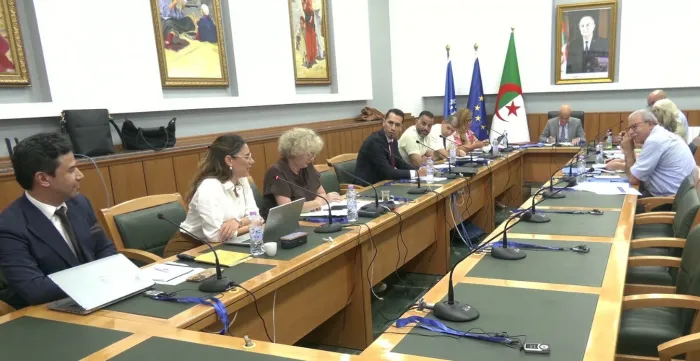 Vue d’ensemble d’une réunion réunissant plusieurs participants autour d’une grande table équipée de microphones et de documents. 